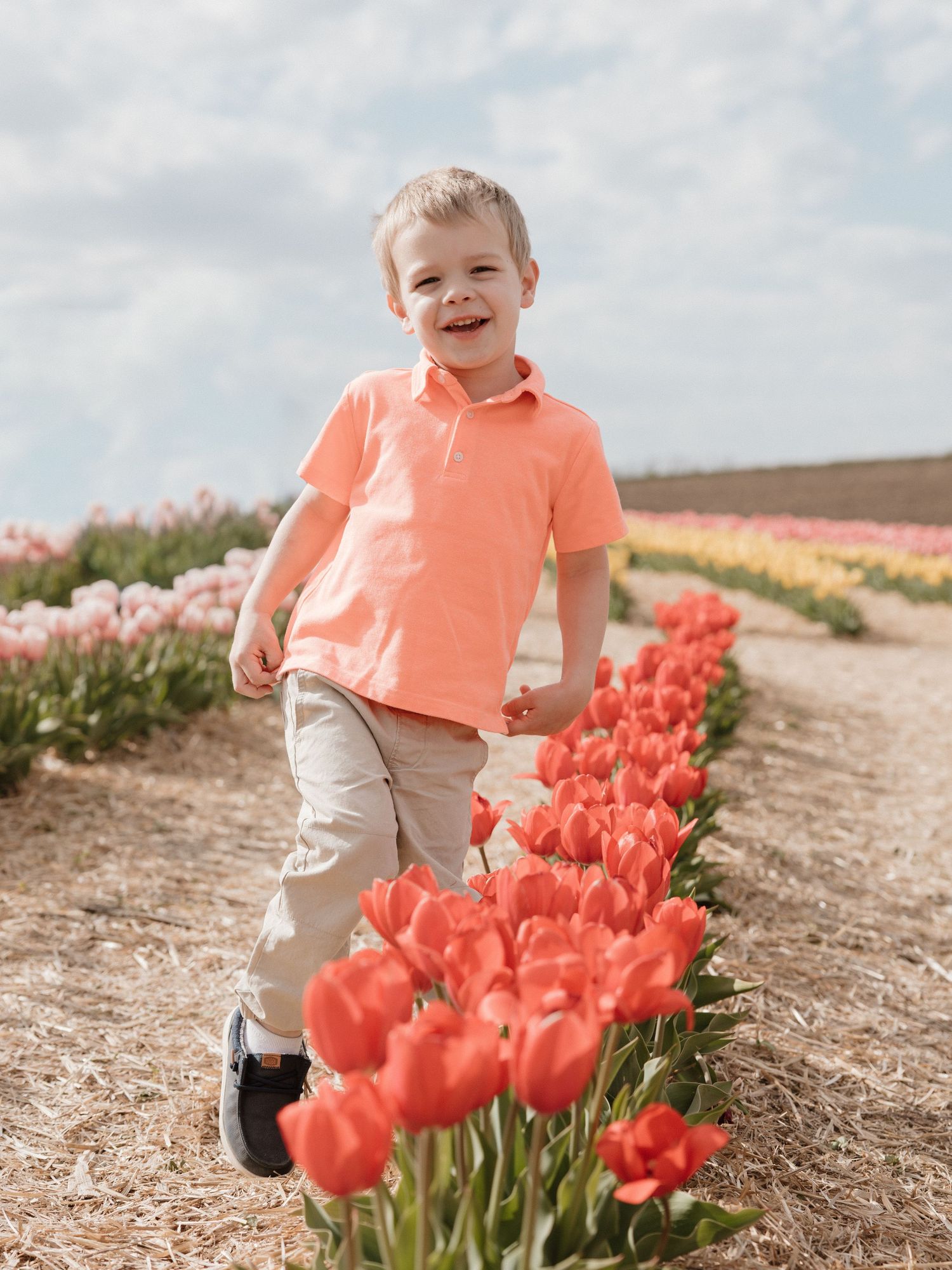 Young boy in coral polo shirt runs joyfully through vibrant tulip fields in bloom on a sunny day.