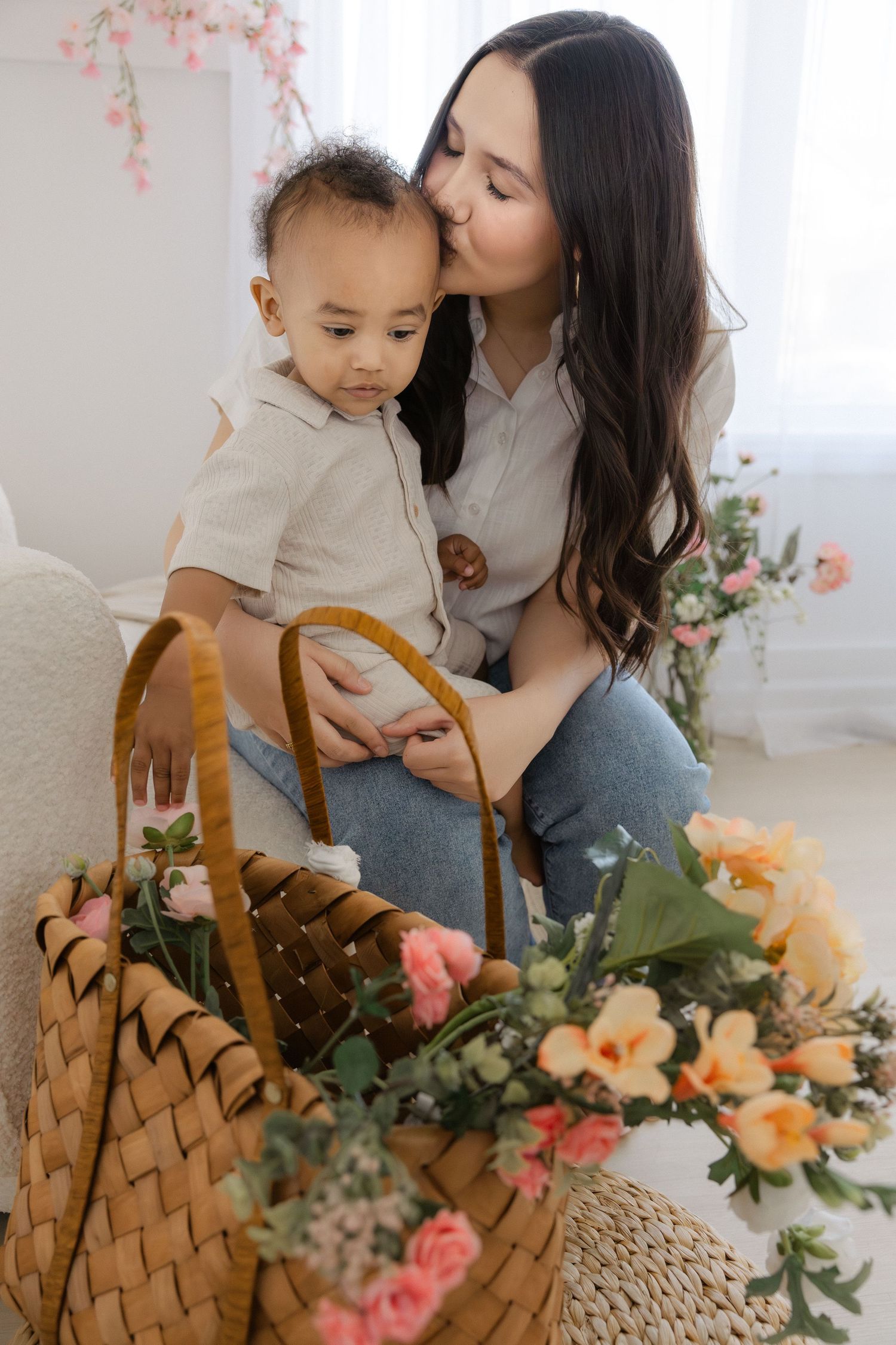 A mother kissing her baby boy while sitting near a wicker basket filled with colorful spring flowers.