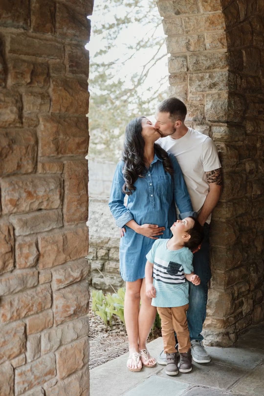 Pregnant woman and man kissing under stone arch with young child standing between them during family maternity photo session.
