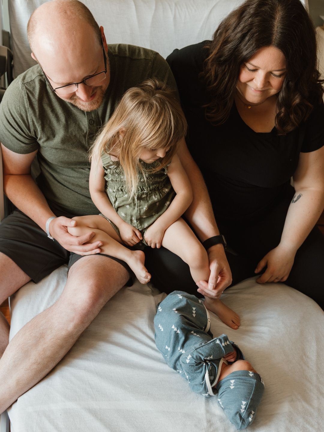 A family of four sits together in a white chair, parents smiling at their toddler and newborn baby wrapped in a gray swaddle.