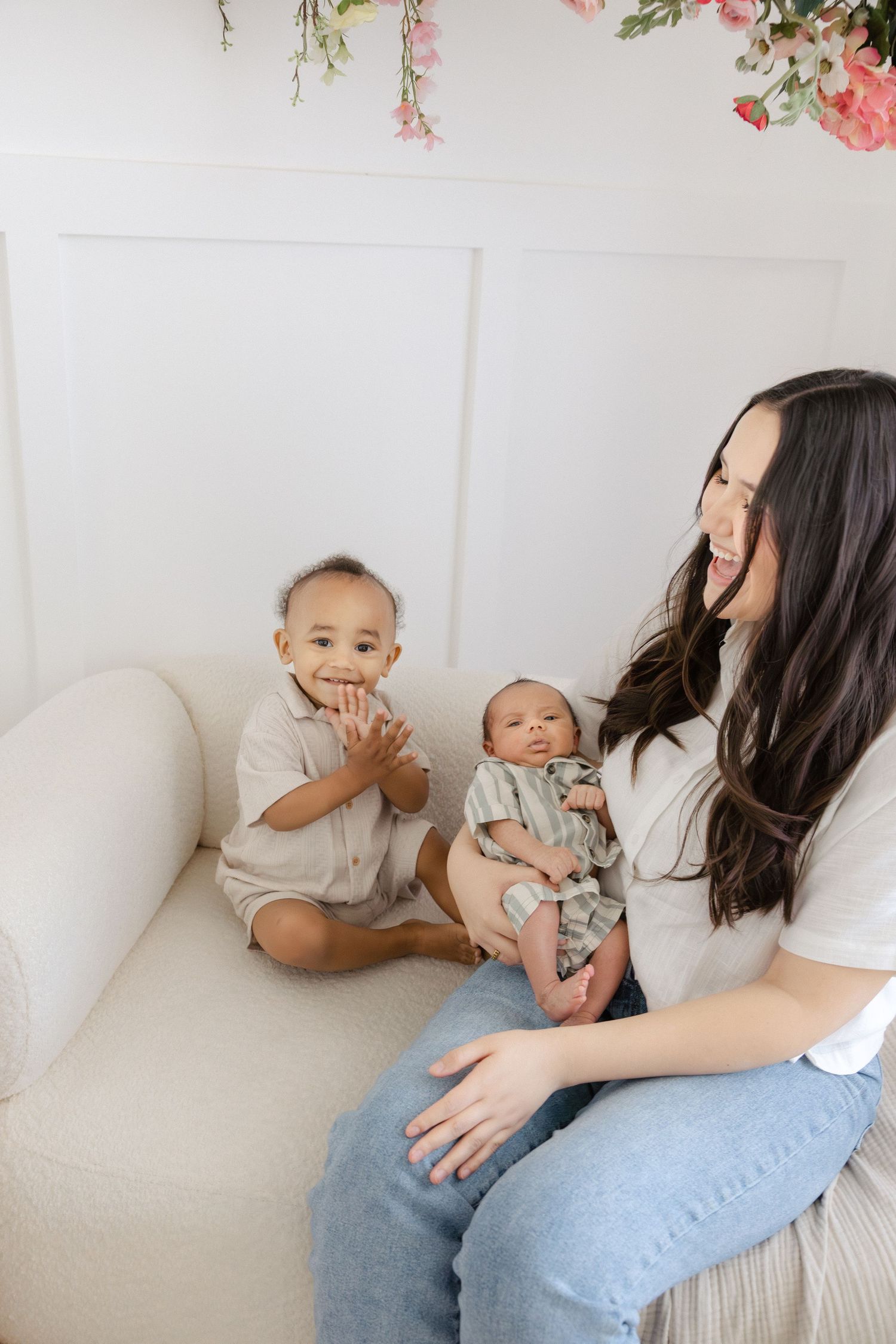 A smiling mother with long dark hair holds a newborn while a toddler sits beside her on a white couch with pink floral decor.
