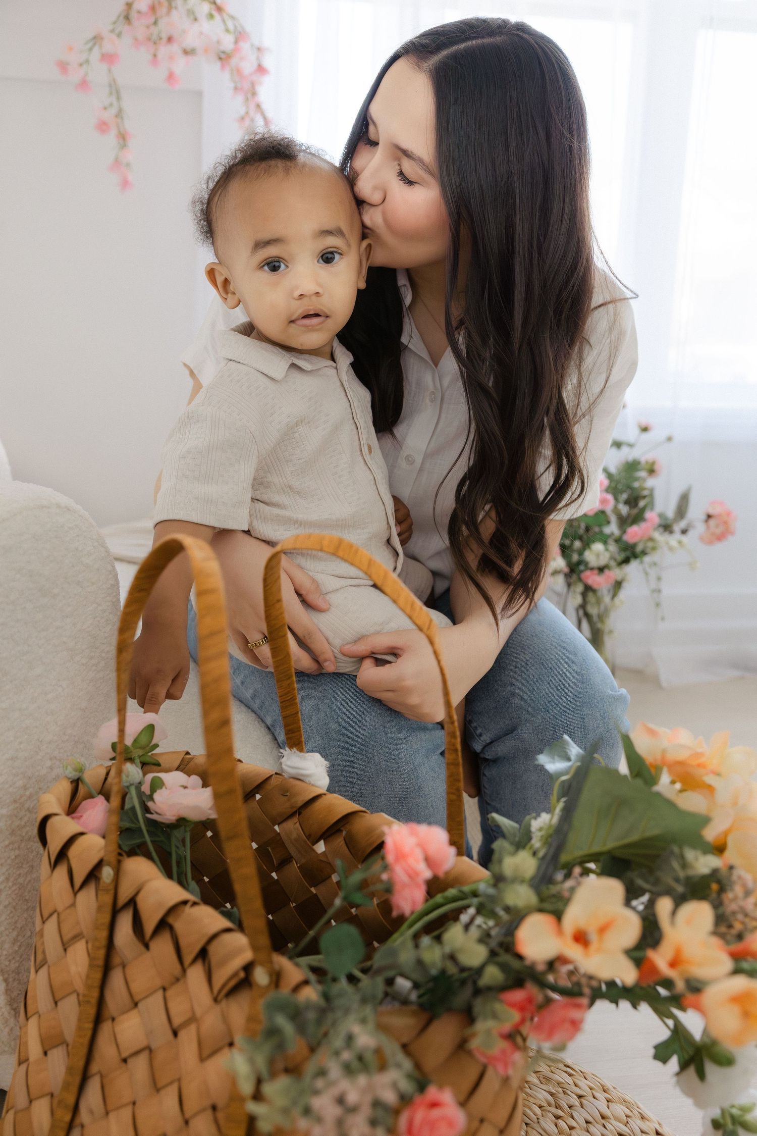A mother kisses her baby boy while sitting near a wicker basket with colorful flowers in a bright room.