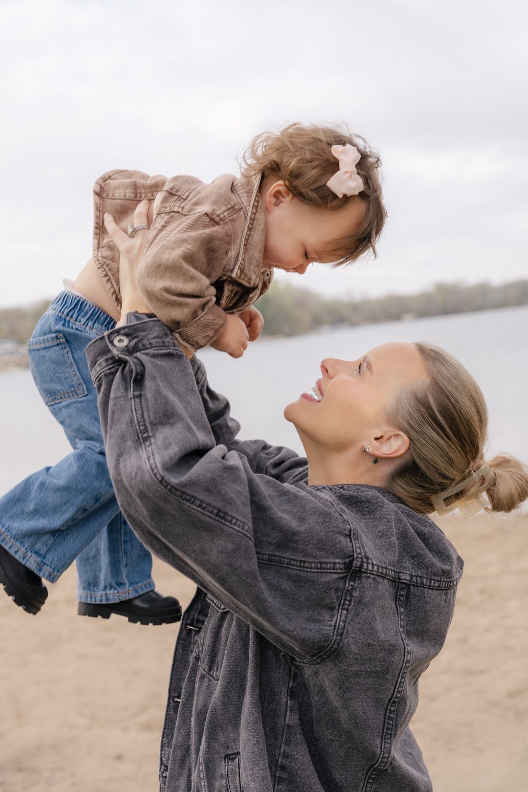 A smiling mother lifts her toddler daughter in the air on a sandy beach near a lake on a cloudy day.