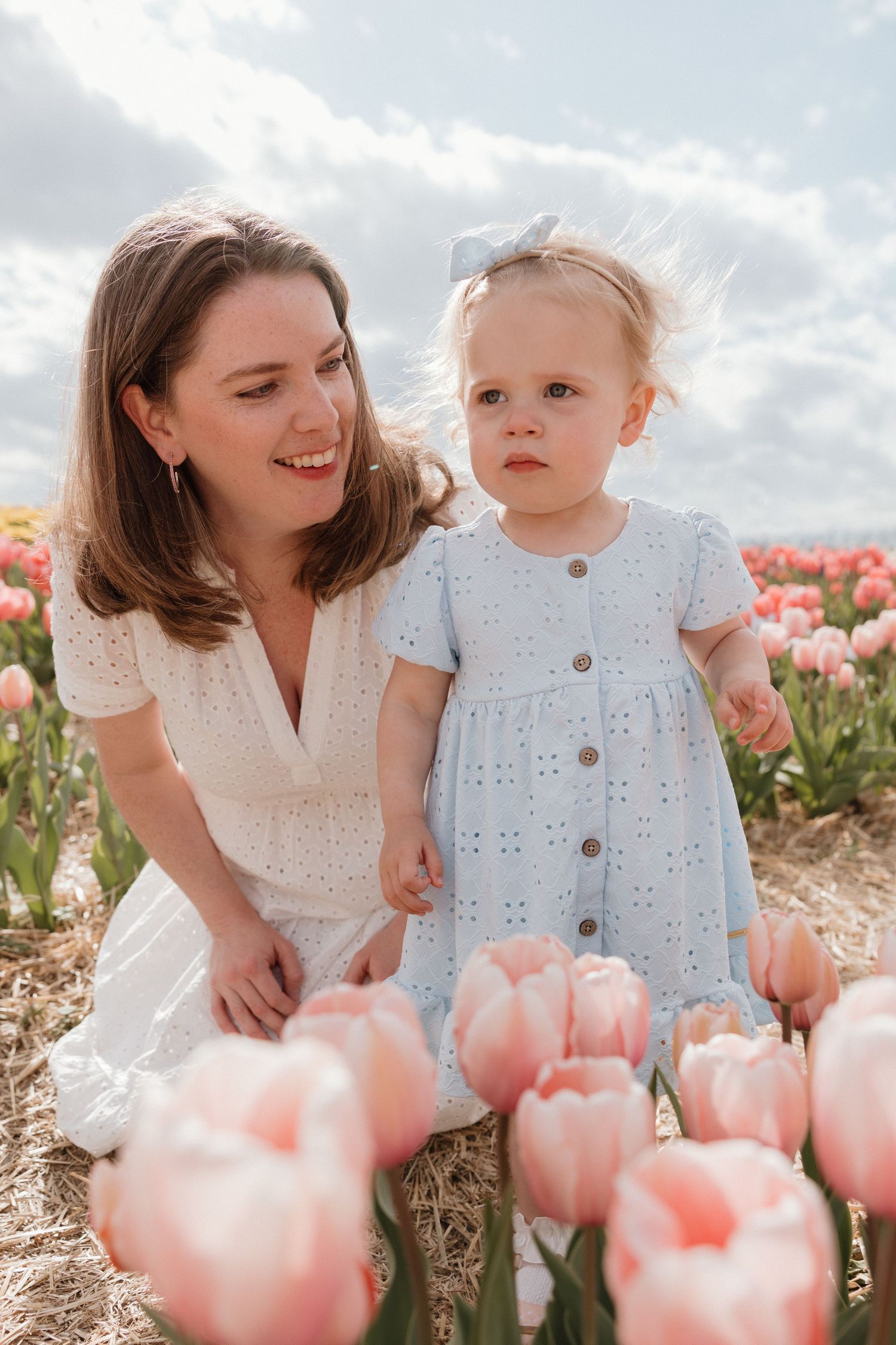 A mother in a white dress smiles at her toddler daughter in a blue dress among pink tulips in a flower field.