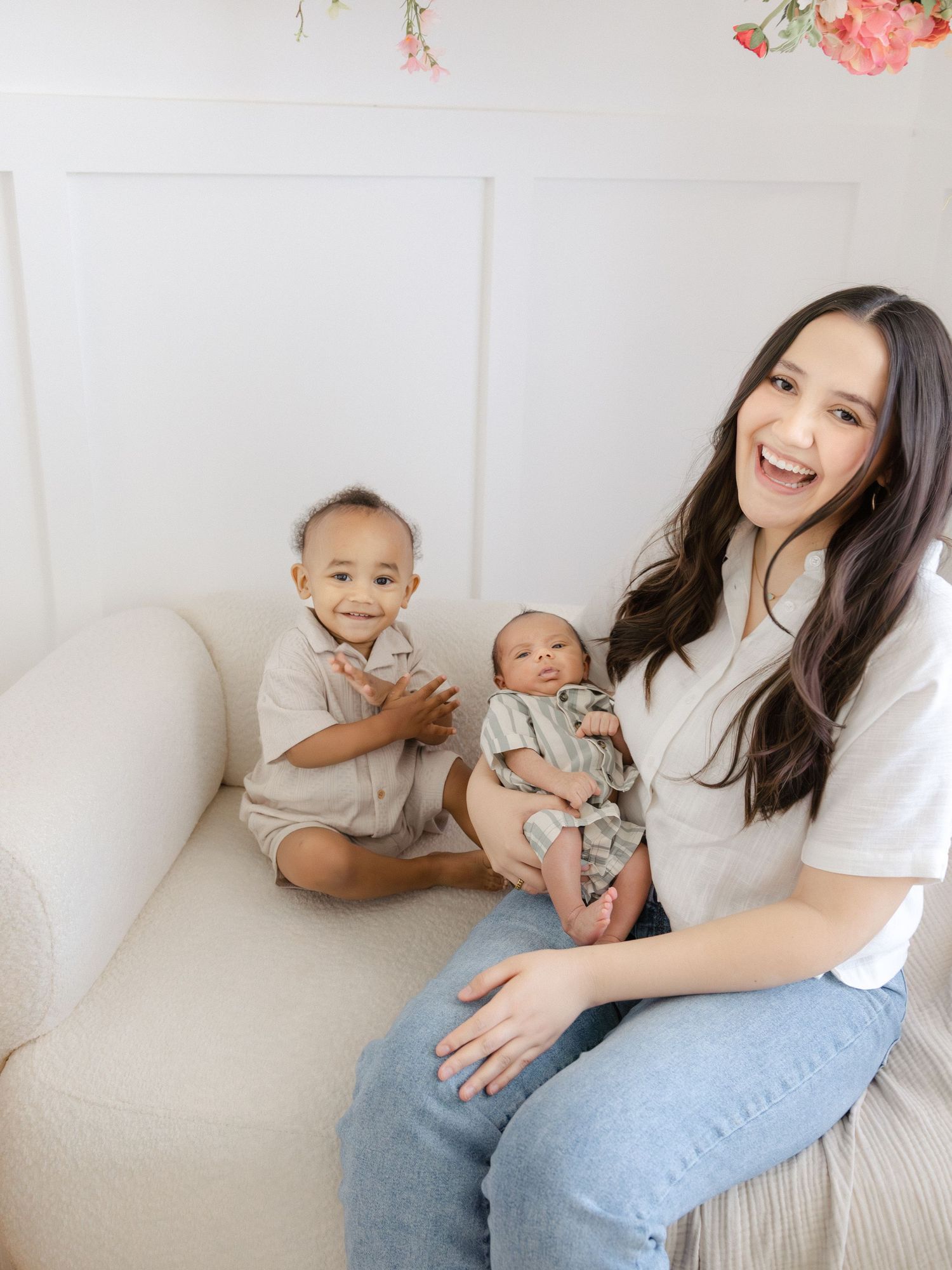 A smiling woman holds a newborn baby while a toddler sits beside her on a cream couch in a bright white studio.