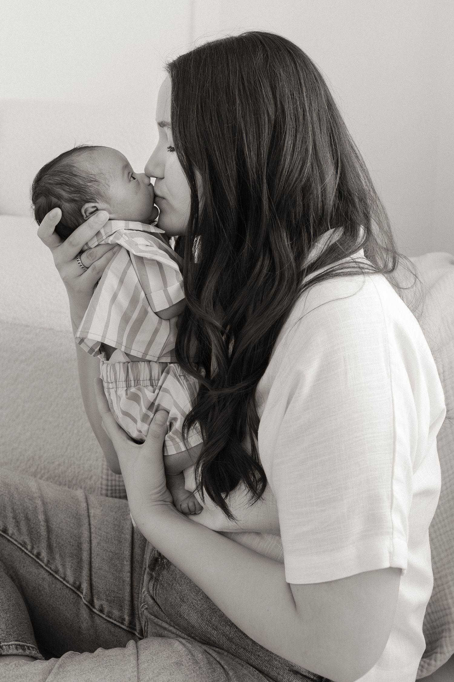 Black and white photo of a mother kissing her newborn baby while sitting on a bed.