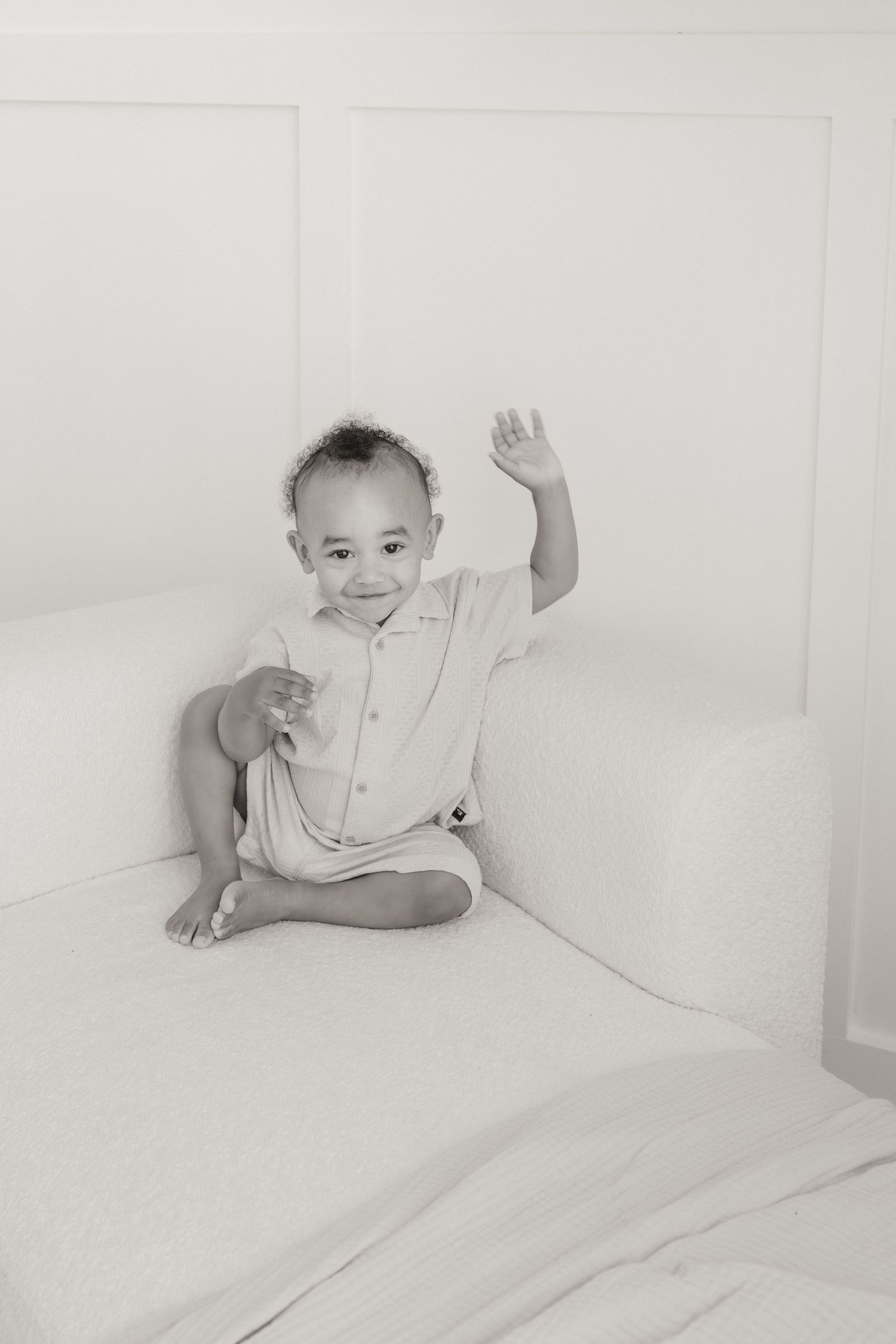 Smiling baby boy sitting cross-legged on white sofa, waving hand, wearing light romper, black and white photo.
