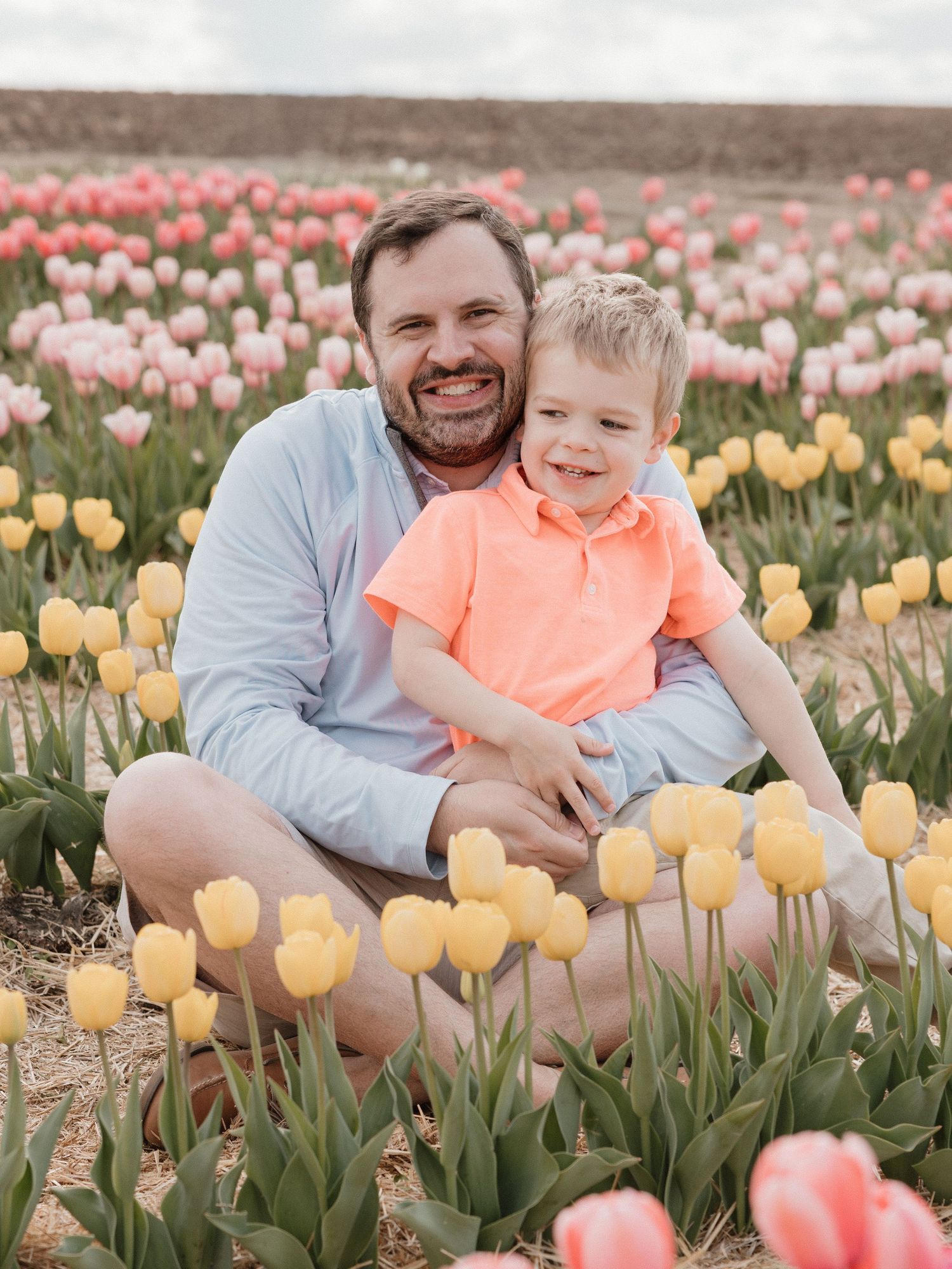 Man and young boy smiling together in a vibrant tulip field with pink and yellow blooms in spring.