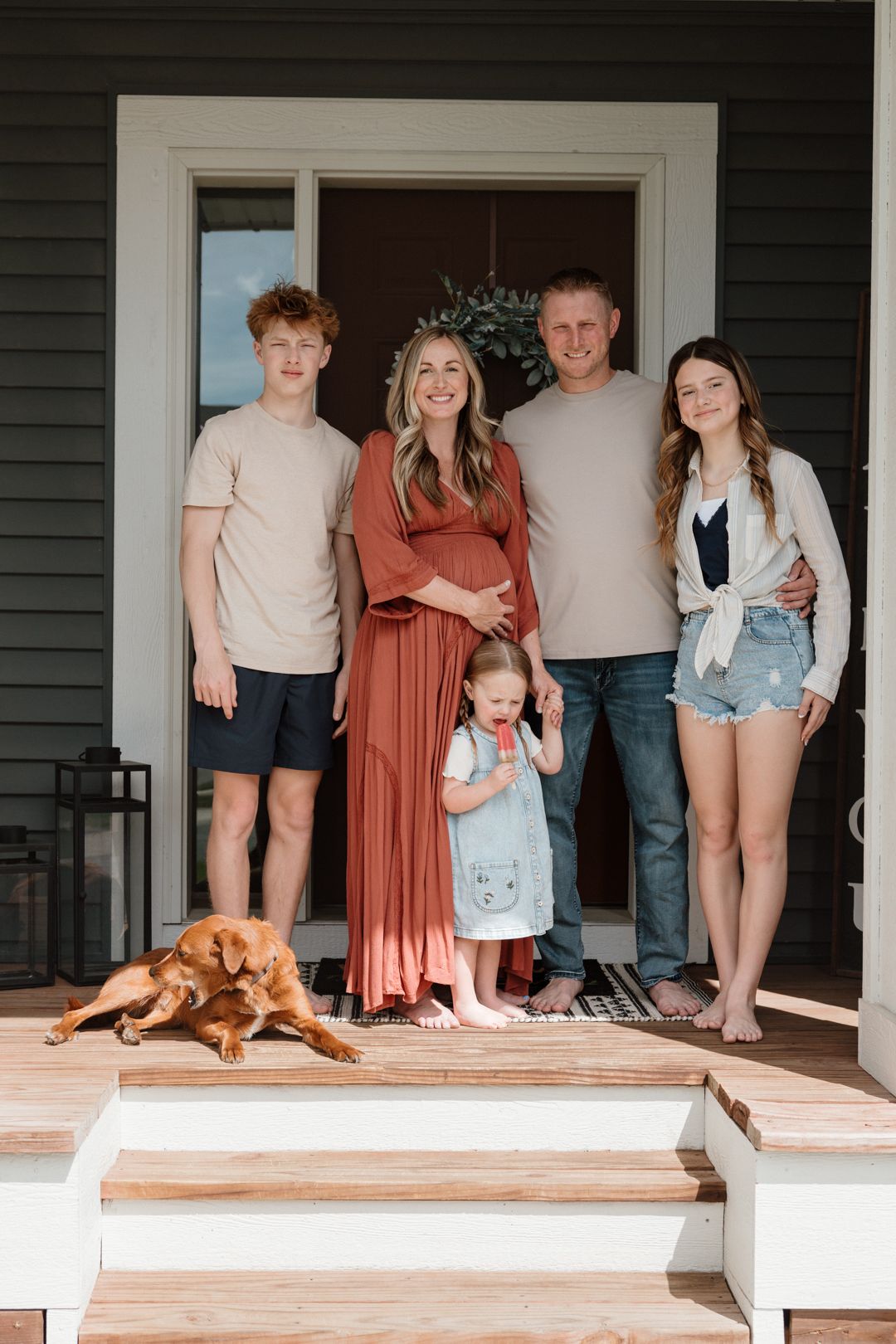 A pregnant woman poses with her family and dog on their home porch for a maternity photo session.