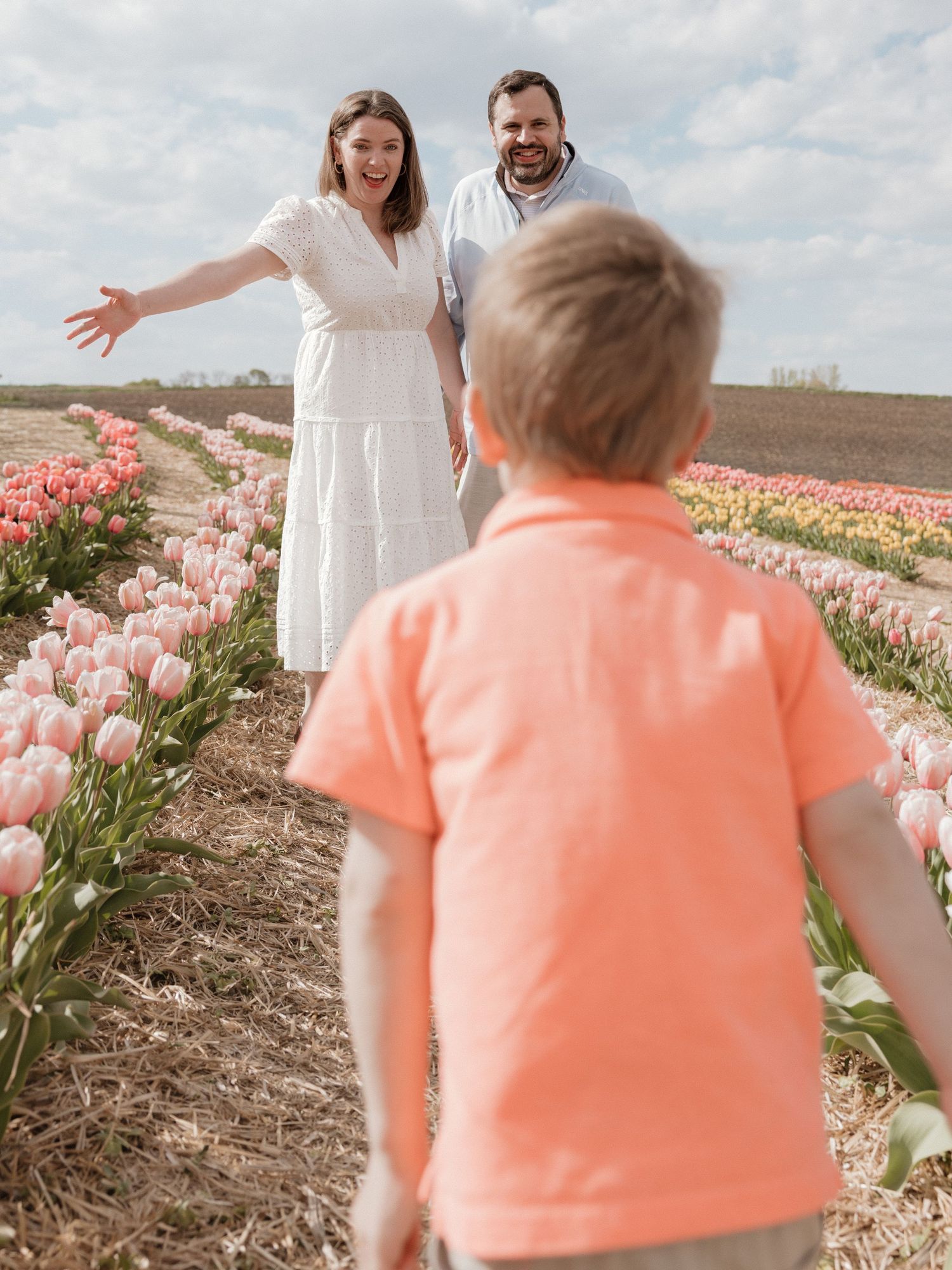 A young boy runs toward his smiling parents standing in a vibrant pink tulip field on a sunny day.