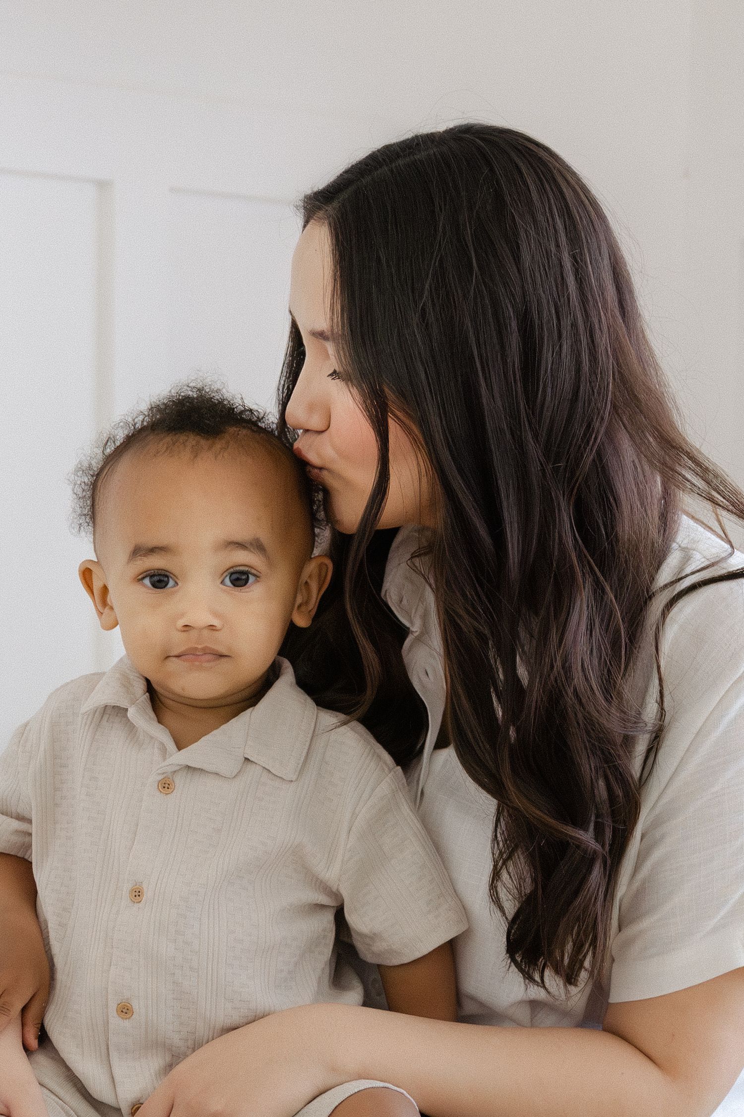 A woman with long dark hair gently kisses a toddler boy on the head while he looks at the camera.
