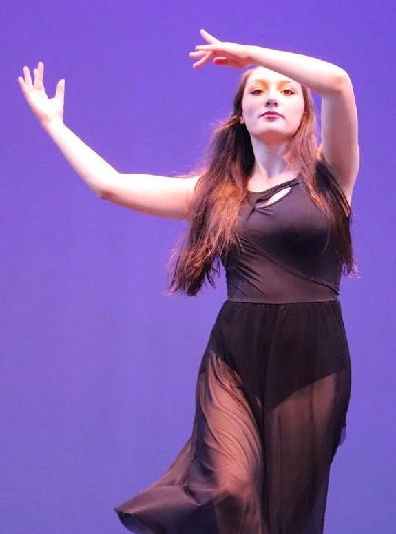 Modern dancer in black flowing costume performing against purple backdrop on stage.