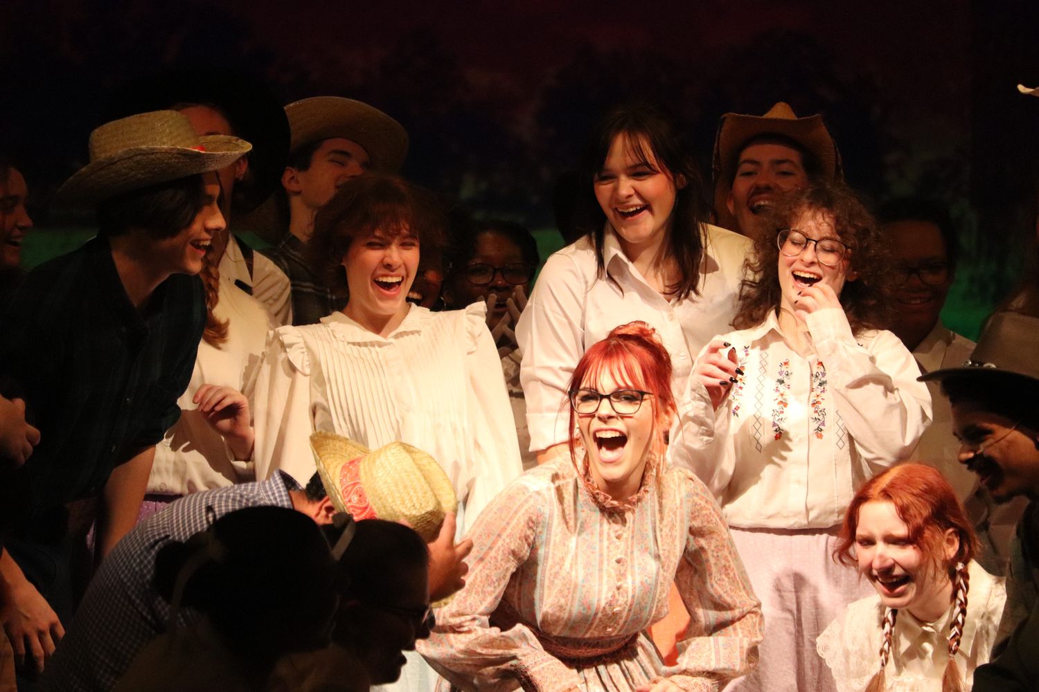 Cast members in period costumes perform an animated scene on a dimly lit stage.