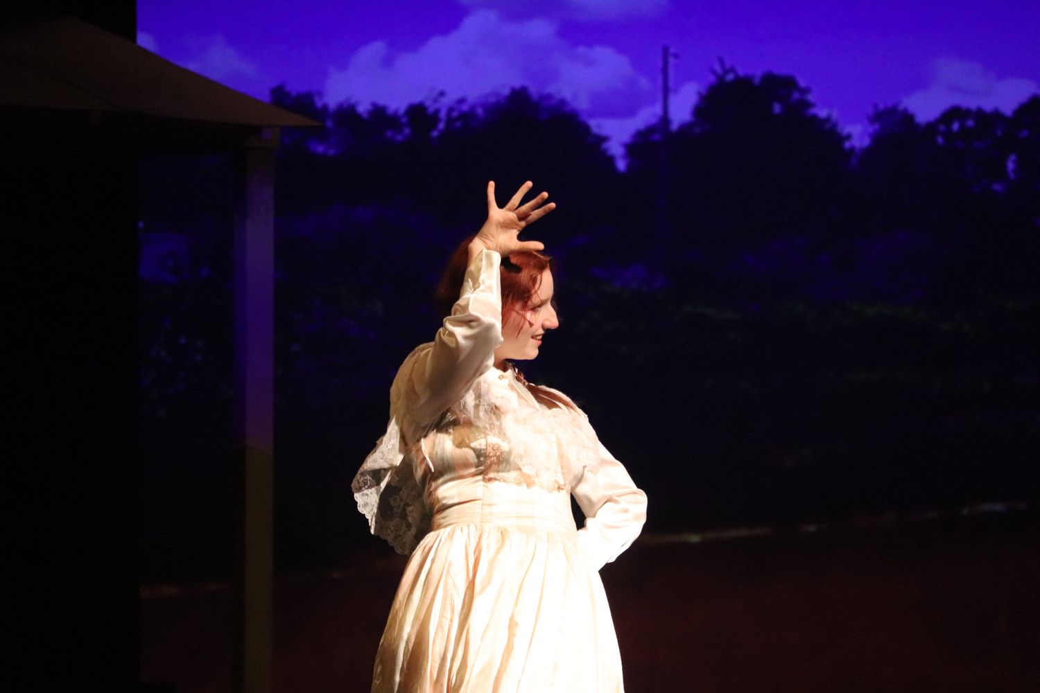 Stage performer in white period dress gestures dramatically against evening sky.