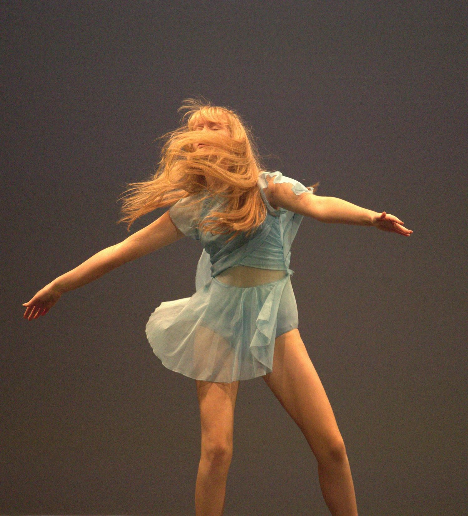 Dancer in light blue dress performs expressive modern dance movements against dark background.