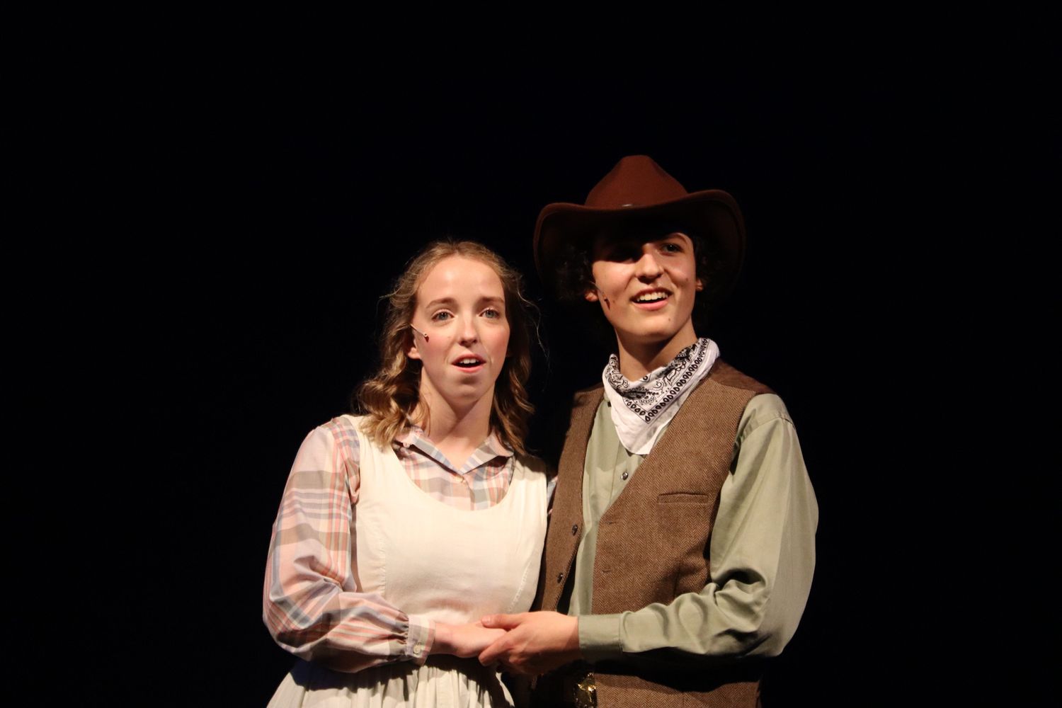 Actors perform scene in period costumes against black background.