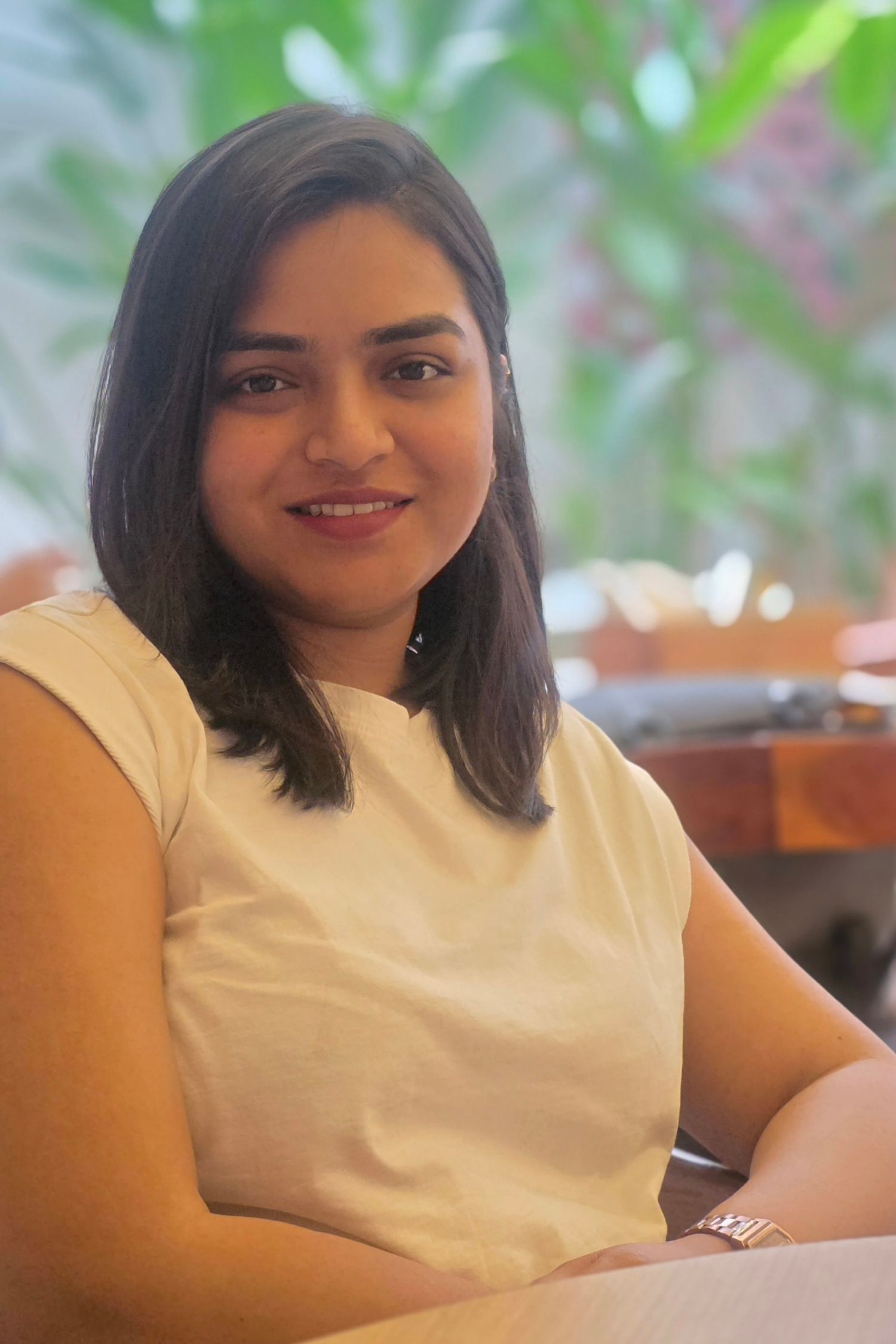 Person in a white top smiles warmly while sitting in front of a blurred green background. Shivani Patil