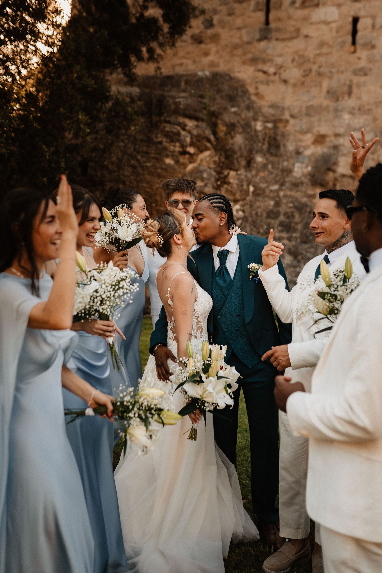 A newly married couple share a kiss while surrounded by their joyful wedding party. Bridesmaids in light blue dresses and groomsmen in white