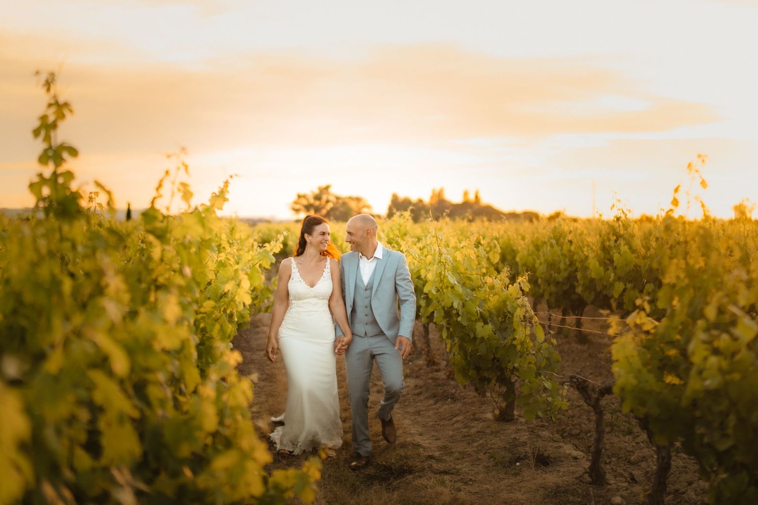 Mariés marchant dans les vignes à la golden hour en Occitanie, lumière chaude et naturelle.