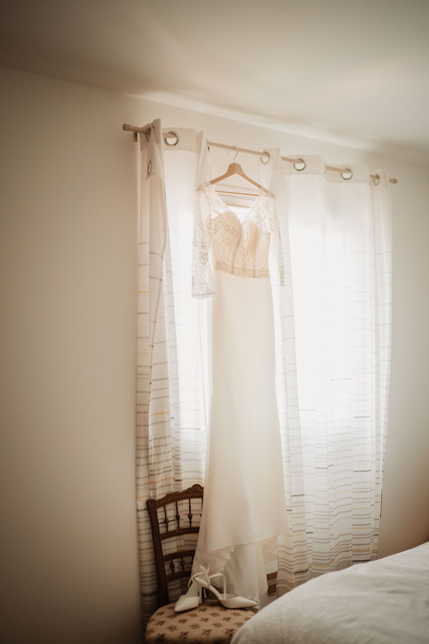 Robe de mariée suspendue devant une fenêtre, lumière douce du matin.
