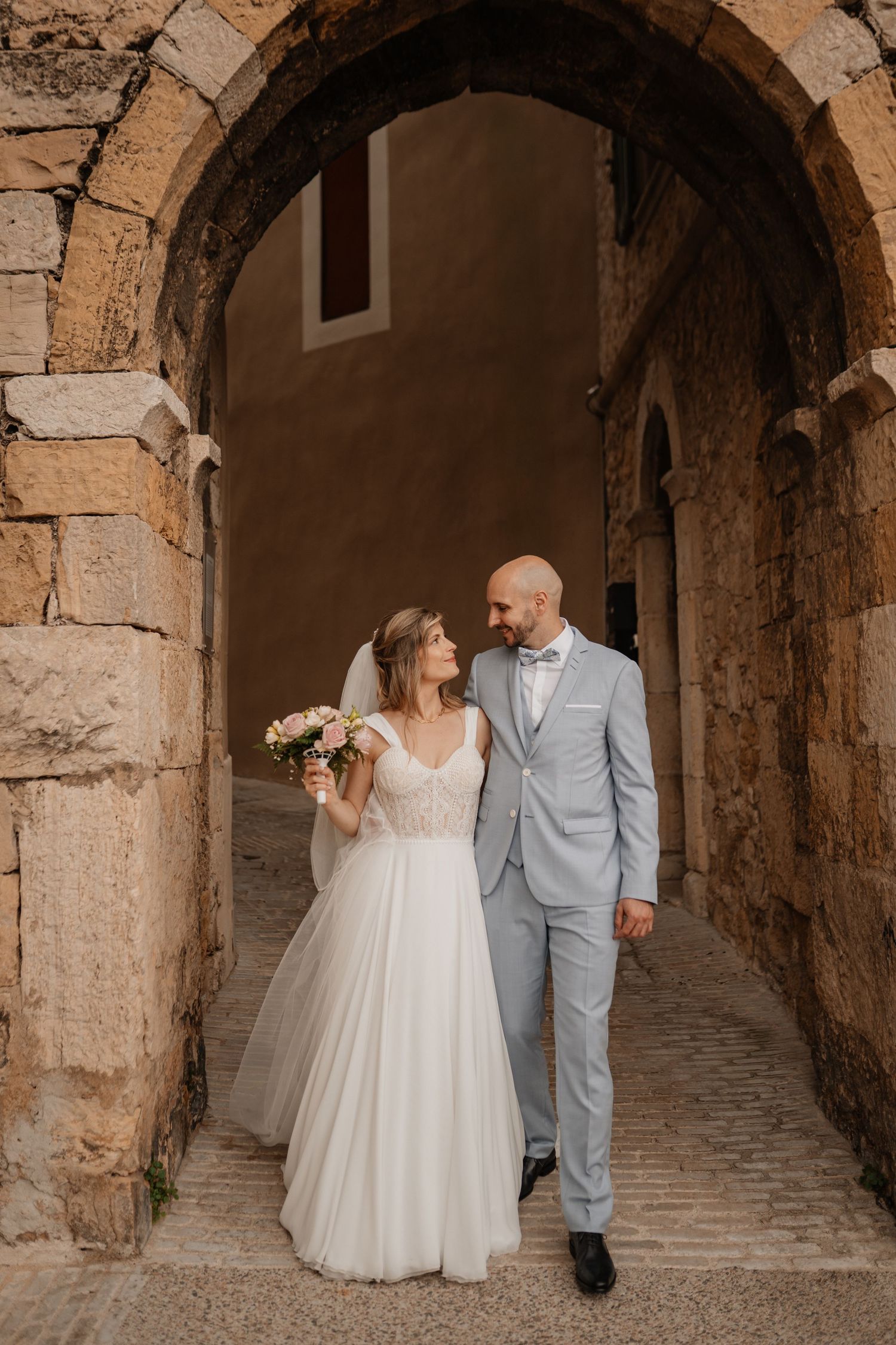 Couple de mariés marchant sous une arche en pierre, mariage champêtre.
