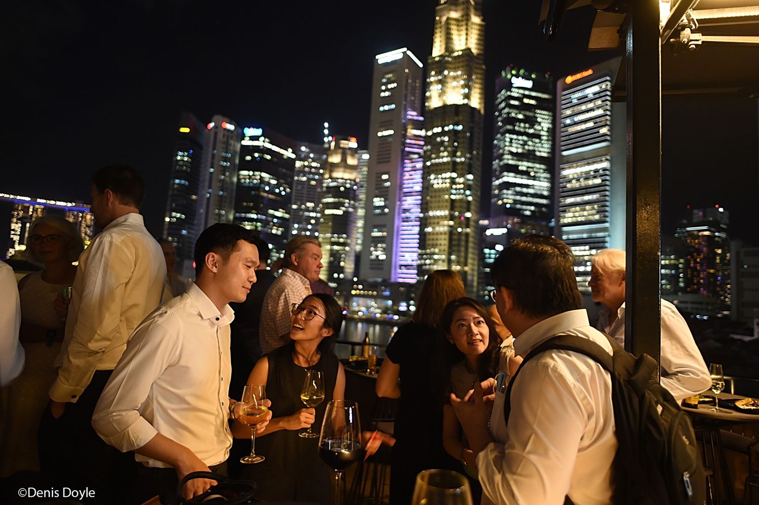 People enjoying drinks at a rooftop bar with illuminated city skyline and skyscrapers in the background at night.