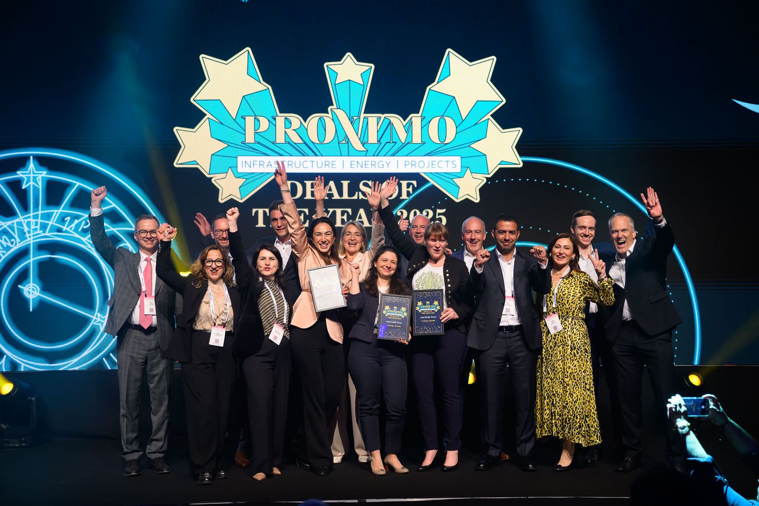 Group of professionals celebrating Pronmo Deals of the Year 2025 award on stage with raised arms and trophy.