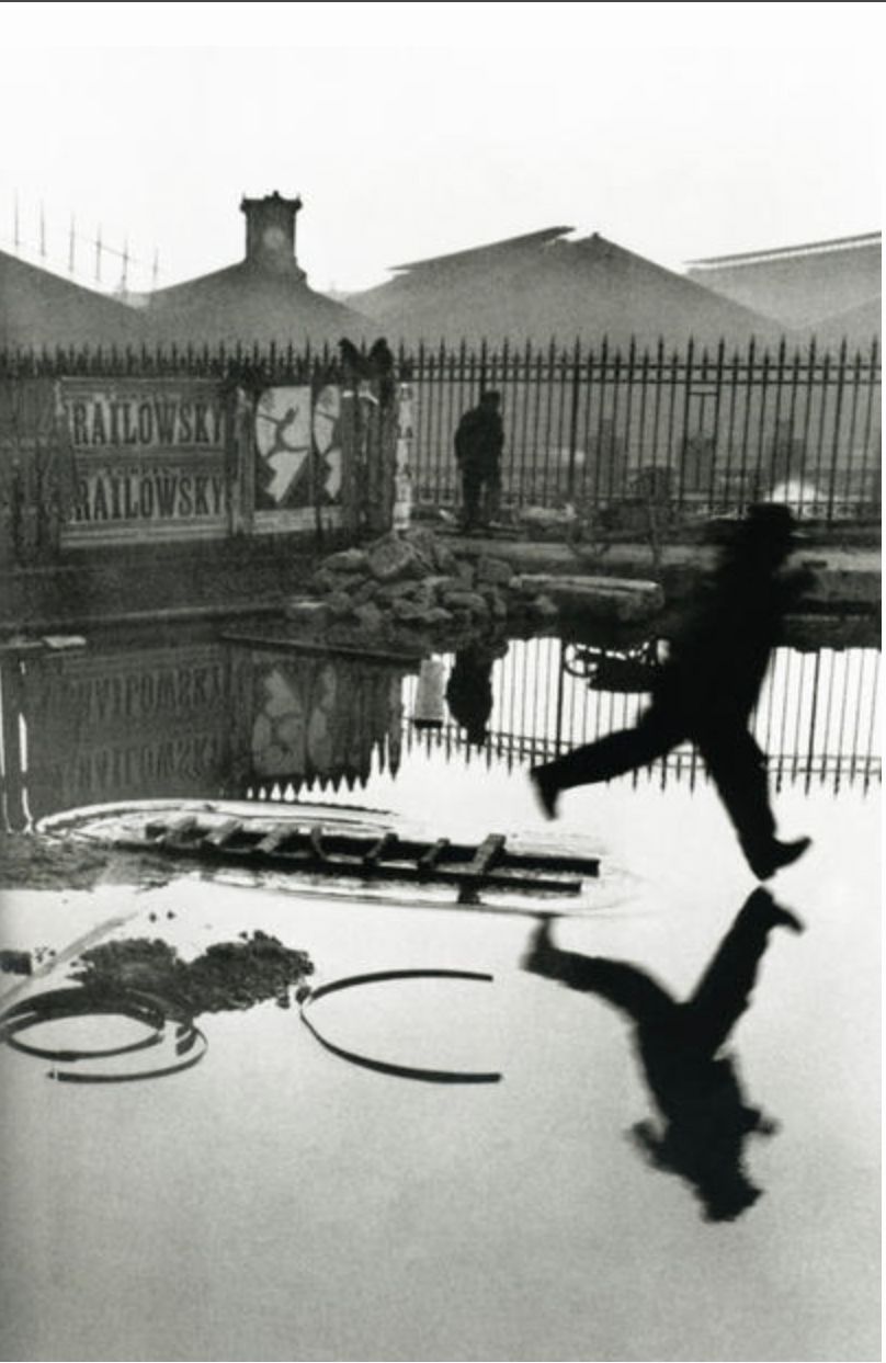 Black and white photo of a man leaping over a puddle near a fence, reflecting posters, in classic street photography style.