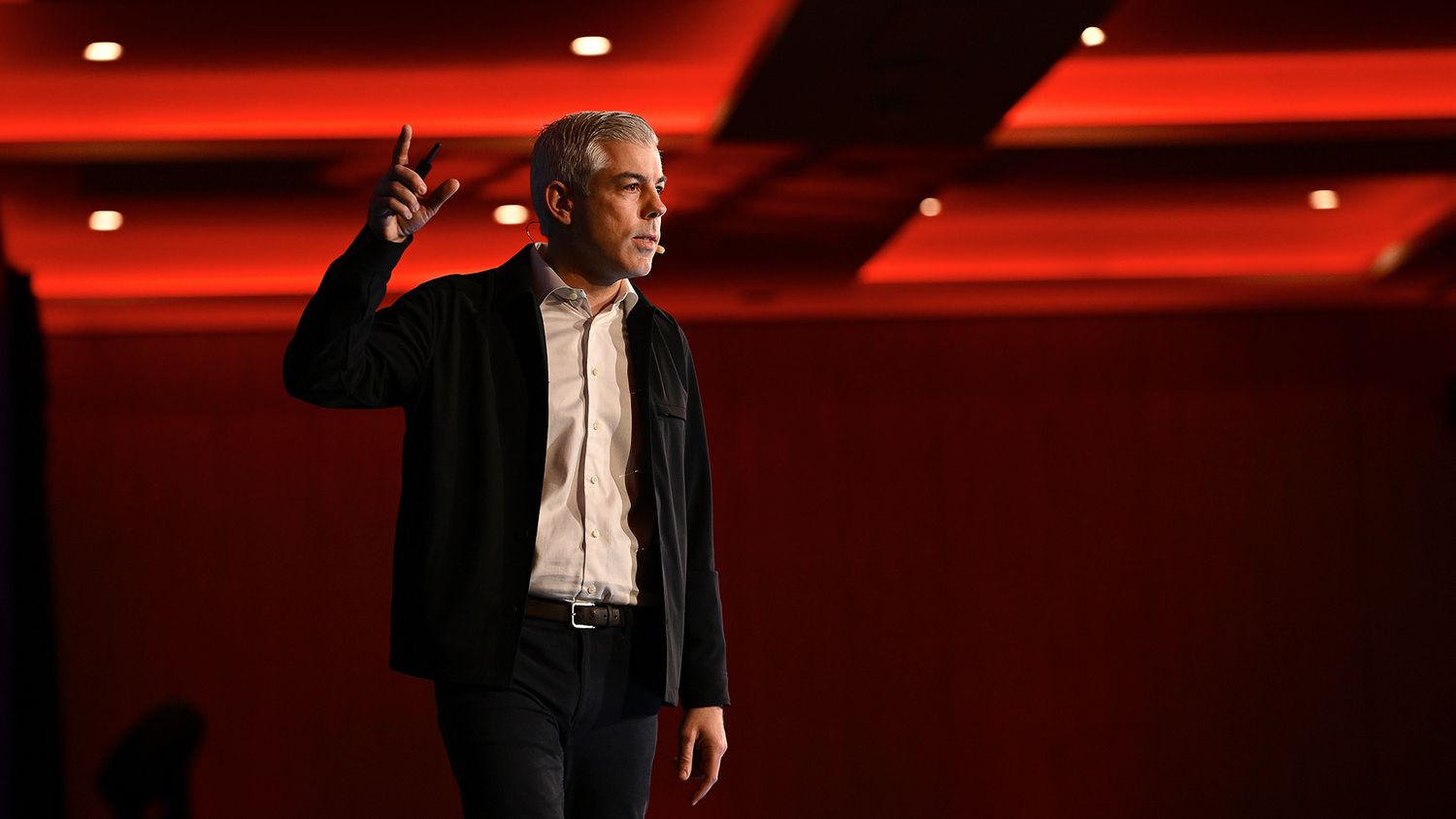 Man in black blazer gestures on a red-lit stage during a professional speaking event or conference.