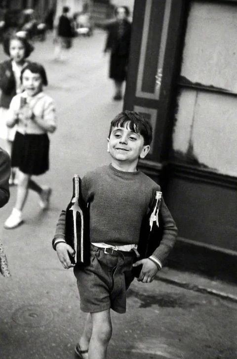 Black and white photo of a smiling boy carrying wine bottles on a cobblestone street in vintage Paris.