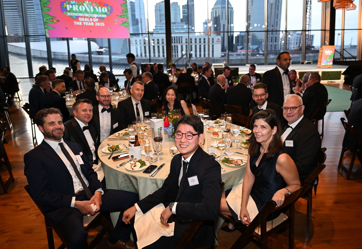 Professionals in formal attire seated at round tables during an awards gala dinner event with city views.