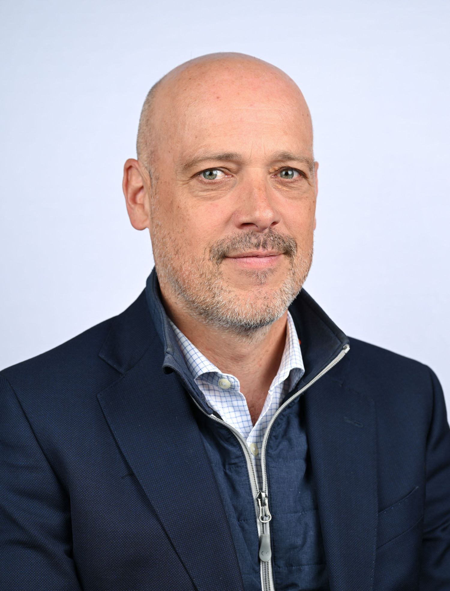 Professional headshot of a bald middle-aged man with a goatee wearing a navy blazer over a zip-up jacket.