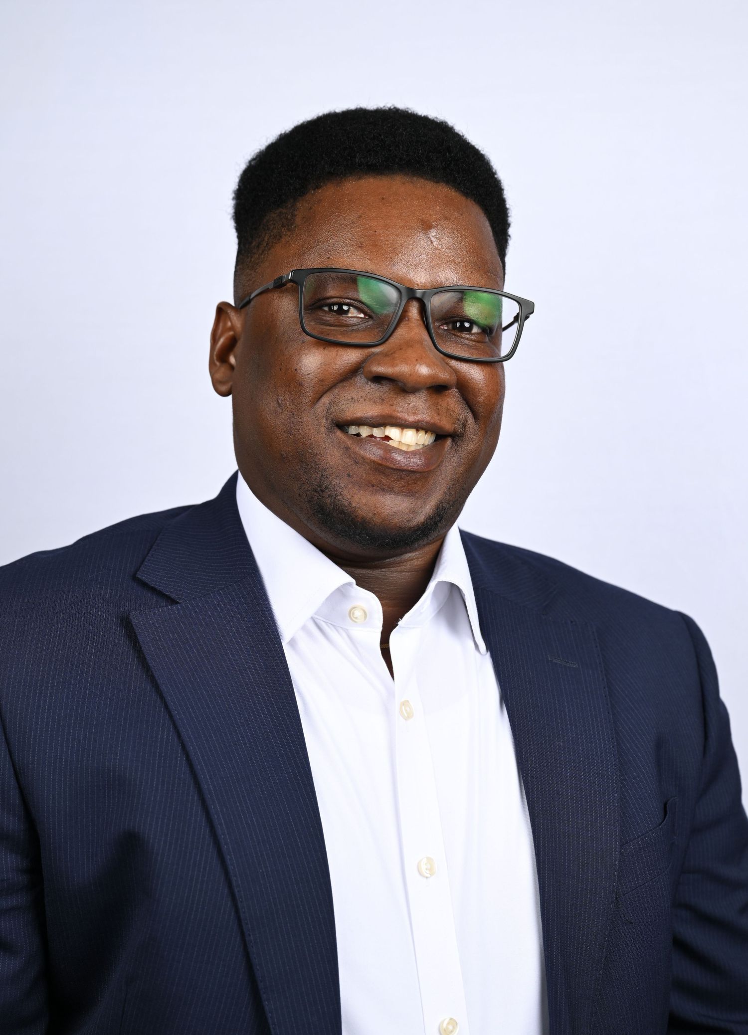 Professional headshot of a smiling man wearing glasses and a navy blazer against a light gray background.
