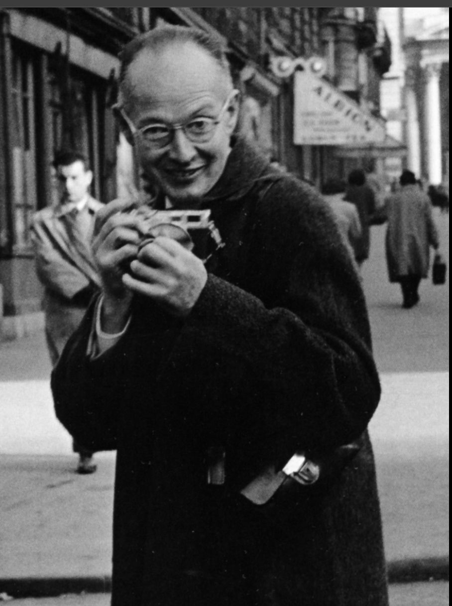 Black and white photo of a smiling man in a coat holding a camera on a busy city street.