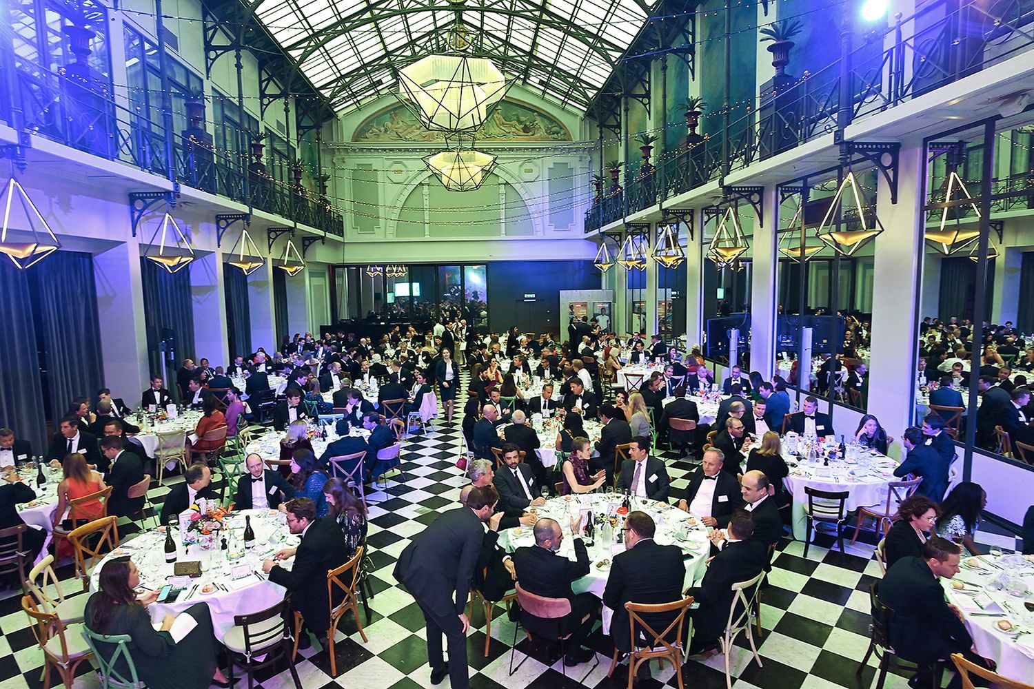 A large, ornate indoor event space with a glass ceiling, filled with tables and people dining and socializing.