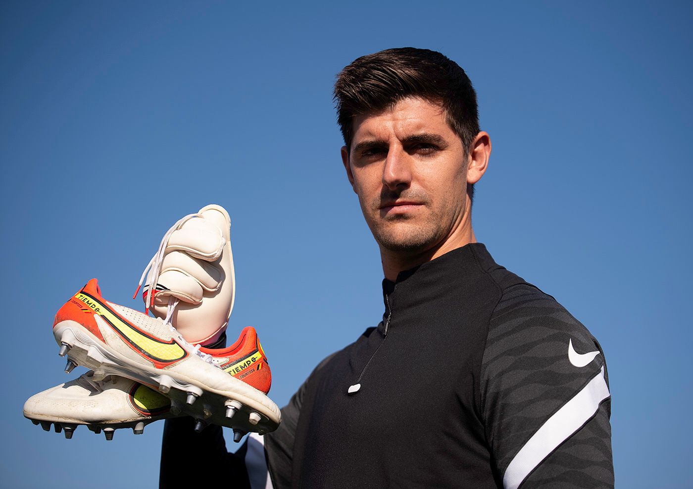 Professional athlete wearing Nike gear poses with soccer cleats against blue sky background.