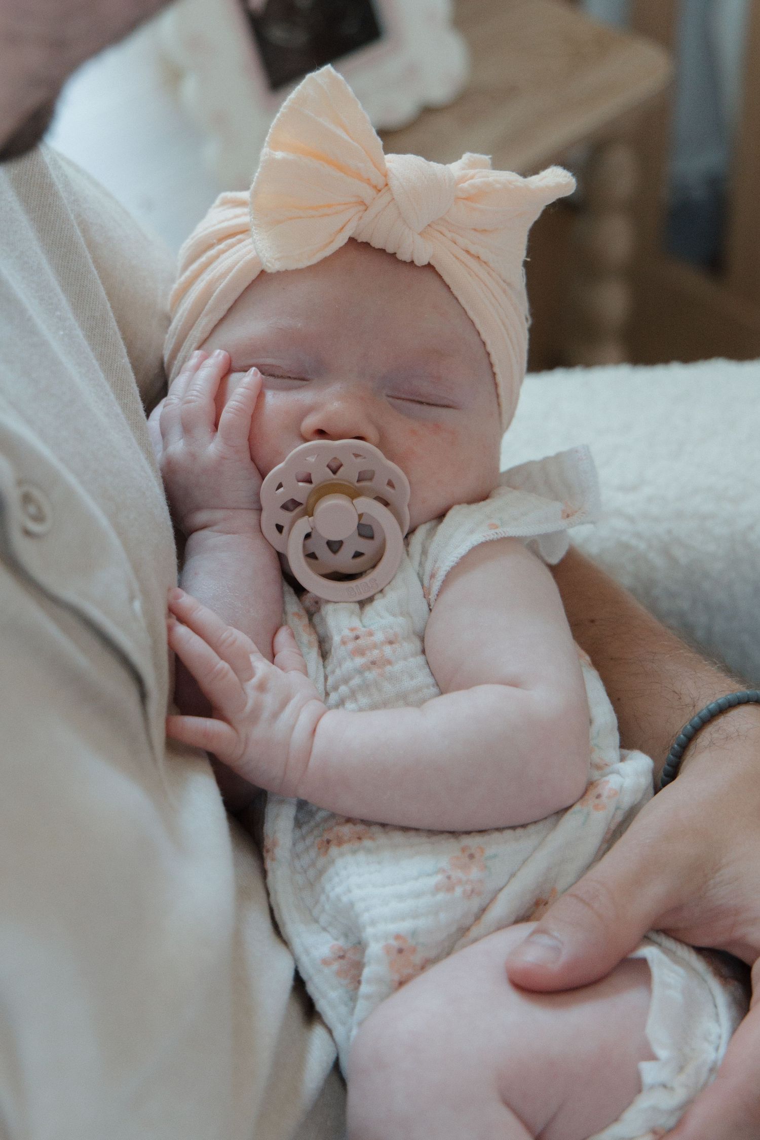 Sleeping newborn baby wearing a peach bow headband and pacifier, cradled in an adult's arms.