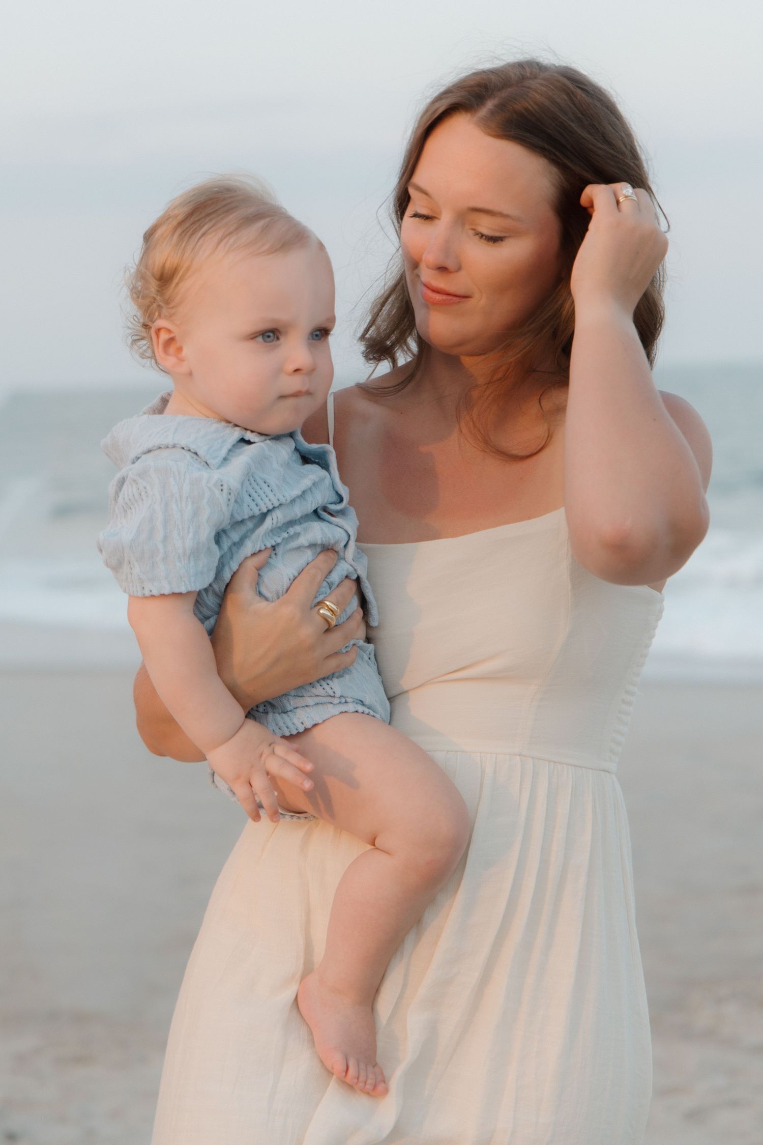 A mother in a white dress holds her baby at the beach during golden hour.