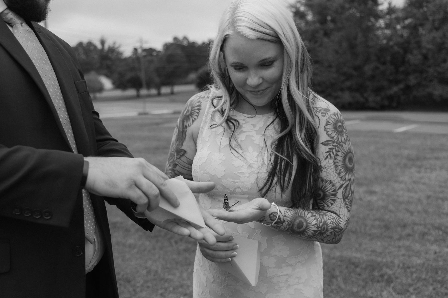 Black and white photo of a tattooed bride in lace dress receiving wedding ring from groom outdoors.