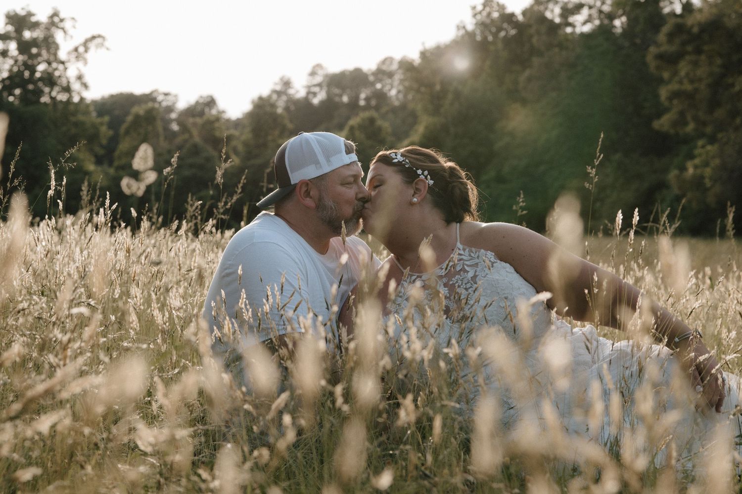 Couple sharing a romantic kiss in a sunlit golden field with tall grass and trees in the background.