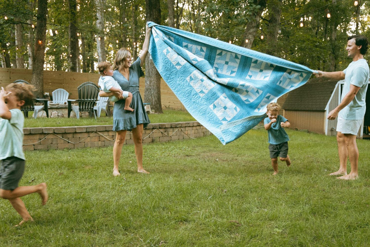 Family playing with a blue quilt blanket in a green backyard surrounded by trees on a warm evening.