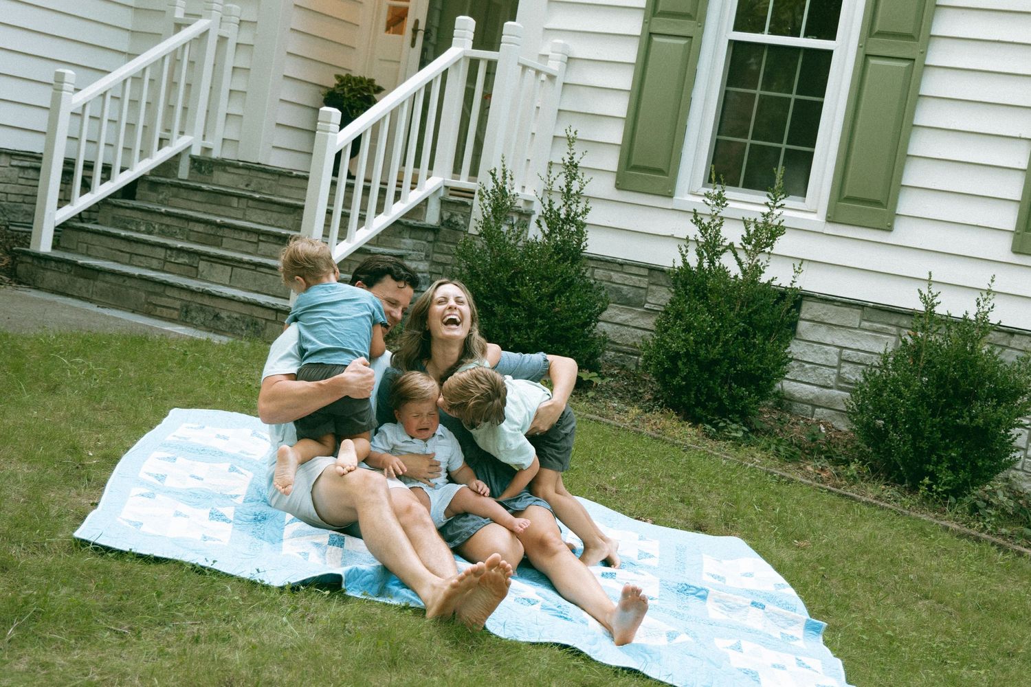 Family laughing and playing together on a blanket in the front yard of a white house with green shutters.