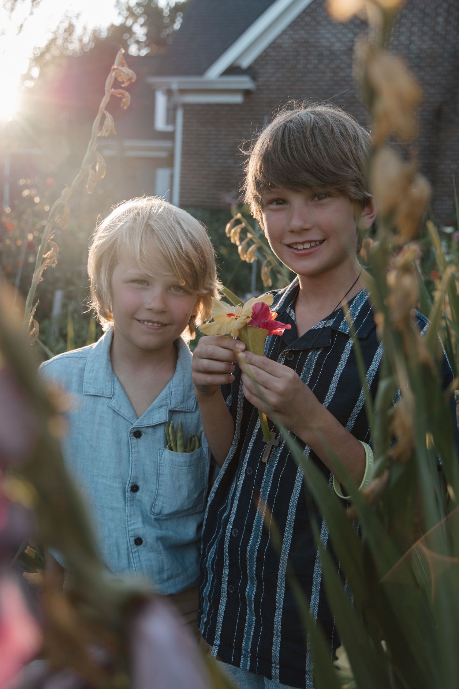 Two smiling boys standing in a sunlit garden holding a yellow and red flower with a brick house in the background.