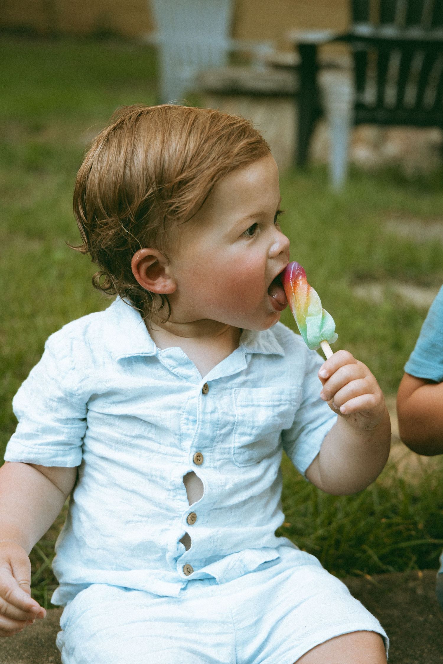 Cute toddler boy in white shirt eating a colorful rainbow popsicle outdoors.