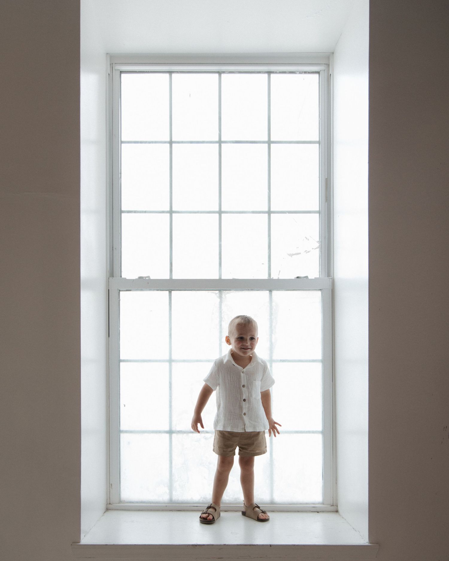 A toddler in a white shirt and tan shorts stands smiling on a windowsill in front of a large white-framed window. CHARLOTTE NC FAMILY