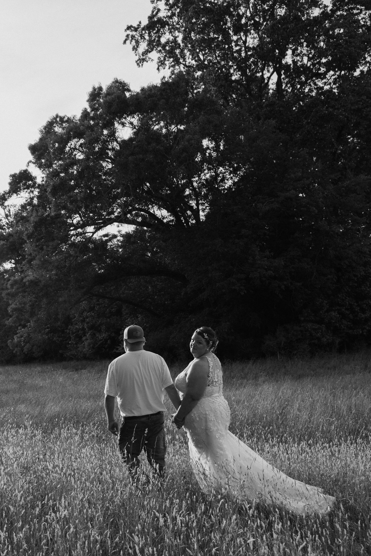 Black and white photo of a bride and groom holding hands walking through a field with large trees in the background.