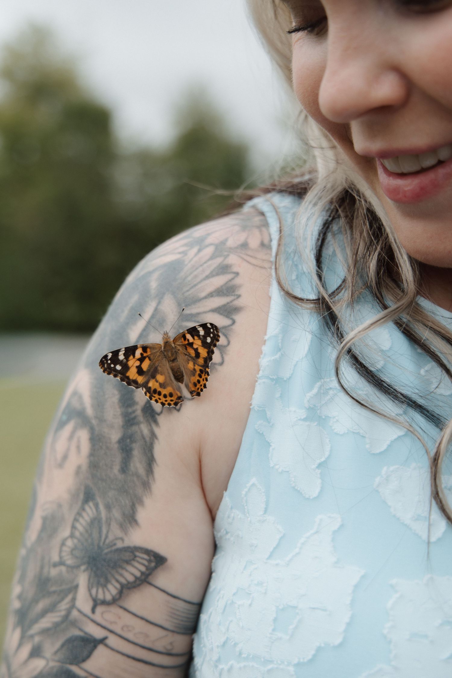 Smiling woman with butterfly tattoo sleeve wearing light blue dress with a painted lady butterfly perched on her shoulder.