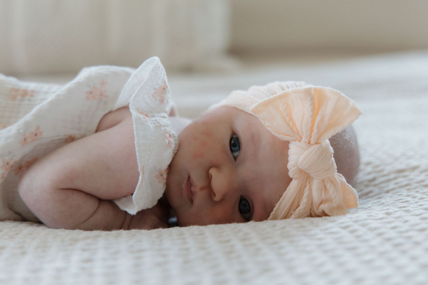 Newborn baby girl in floral outfit and peach bow headband lying on white textured blanket.