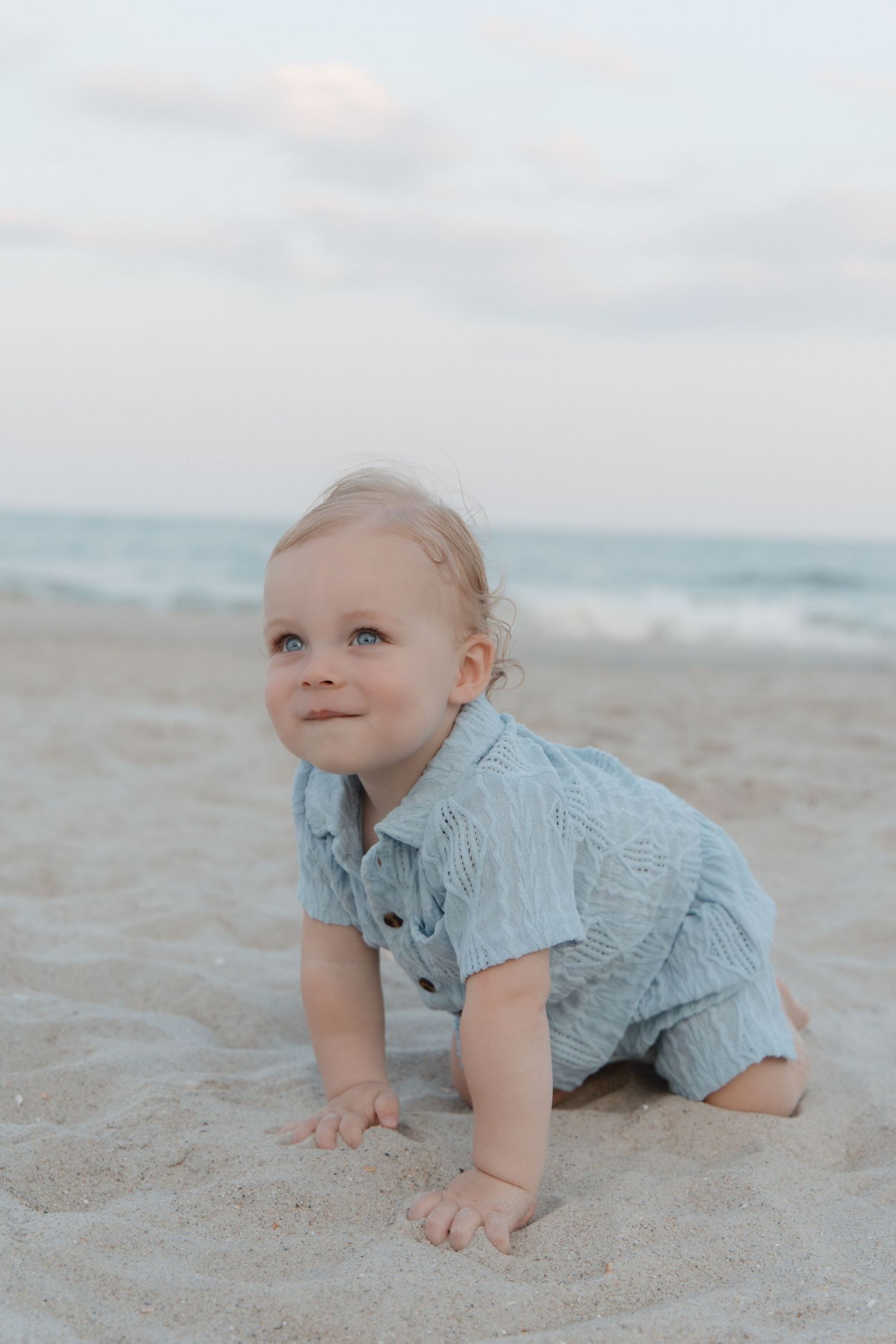 Adorable baby in blue romper crawling on sandy beach with ocean waves in background.