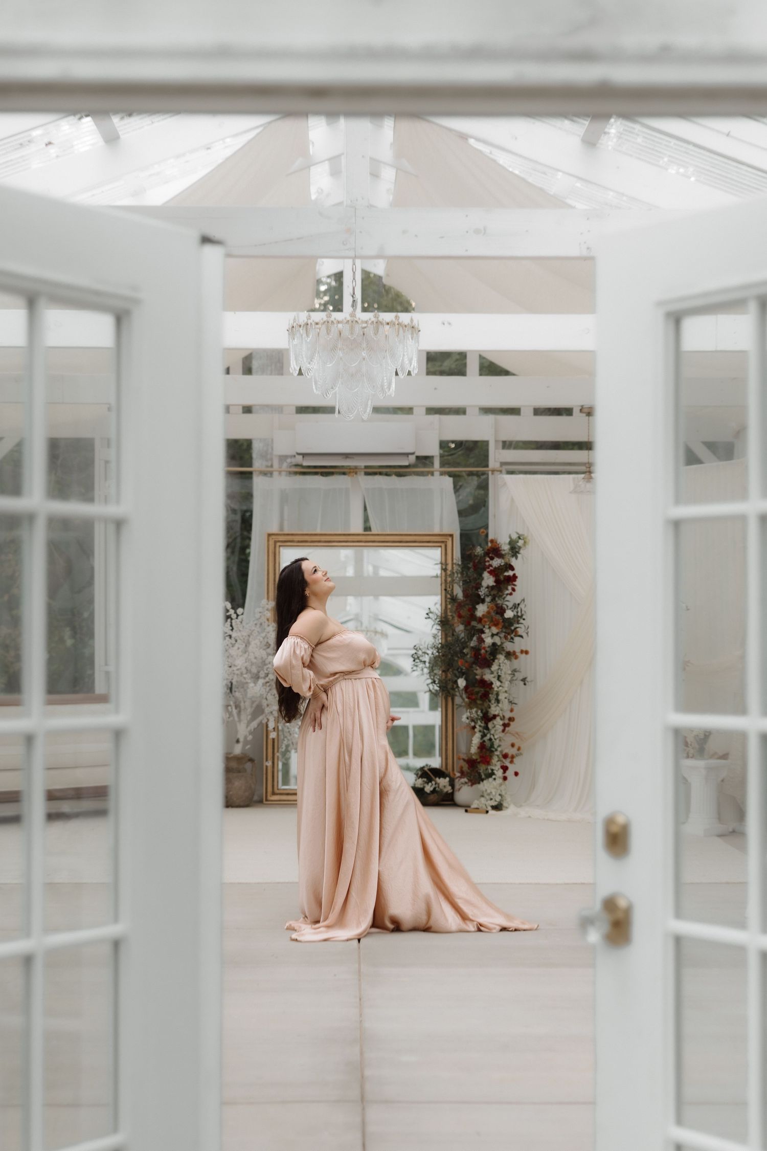 Woman in blush pink gown standing in elegant greenhouse venue with chandelier and floral decor.